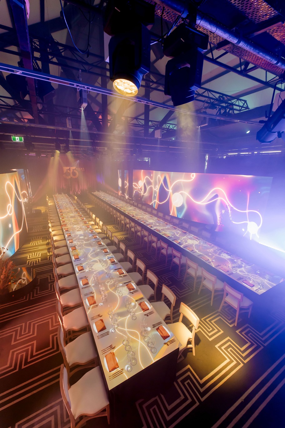 A large, elegant banquet hall with two long dining tables set with glassware and napkins. The room features modern lighting, glowing abstract wall projections, and a high ceiling—perfect for the Doltone 55 Years VIP Qantas Platinum Member Experience.