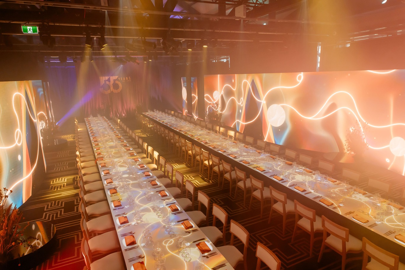 A modern banquet hall with two long dining tables set for an event, decorated with elegant lighting and vibrant orange and gold swirling light projections on the tables and walls.