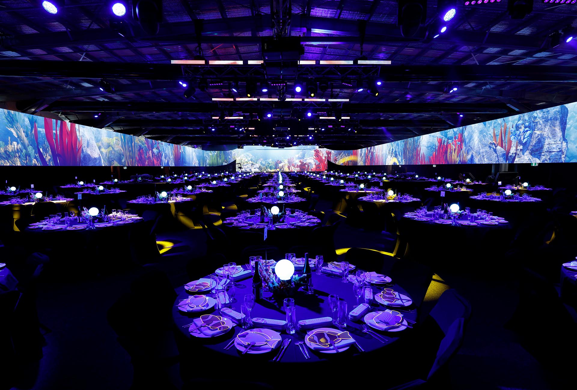 A large event hall set for a formal dinner with round tables, elegant place settings, and glowing centerpieces. The space, designed for a destination sales incentive, features blue and purple lighting with digital screens showing vibrant underwater scenes.