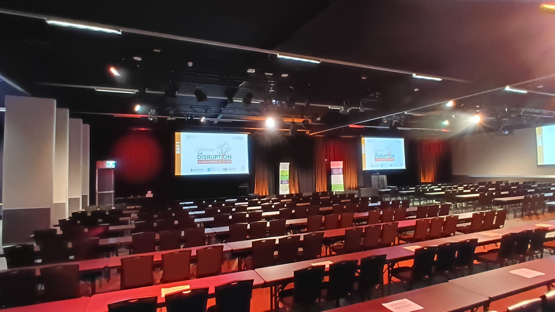 A large conference room with rows of tables and chairs facing three projection screens displaying an event titled Disruption. The room is empty, with stage lighting and banners near the front.
