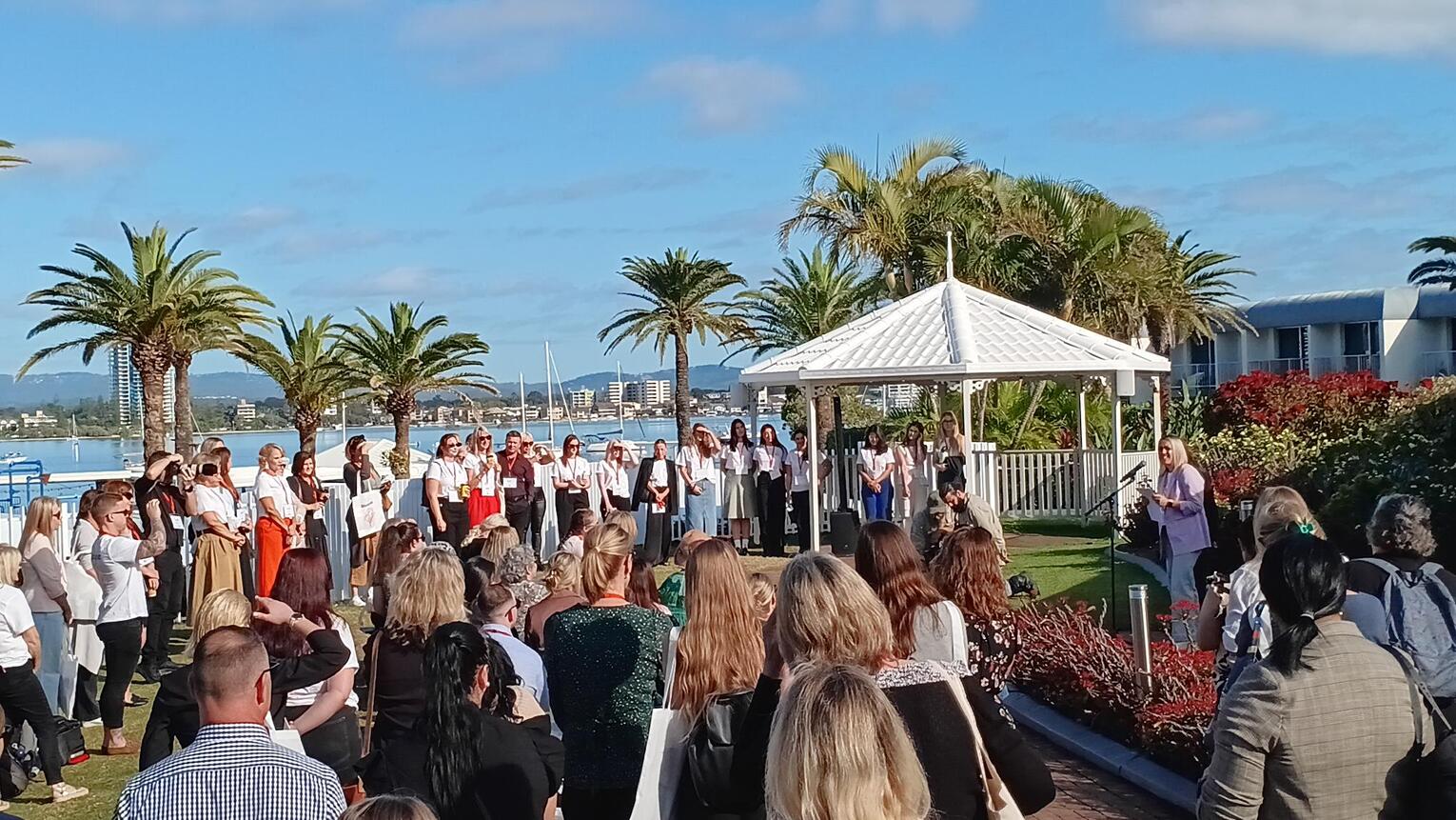 A large group of people gathers outdoors near a white gazebo by the water, surrounded by palm trees and buildings. Some people are standing in a semicircle while one person speaks to the crowd.