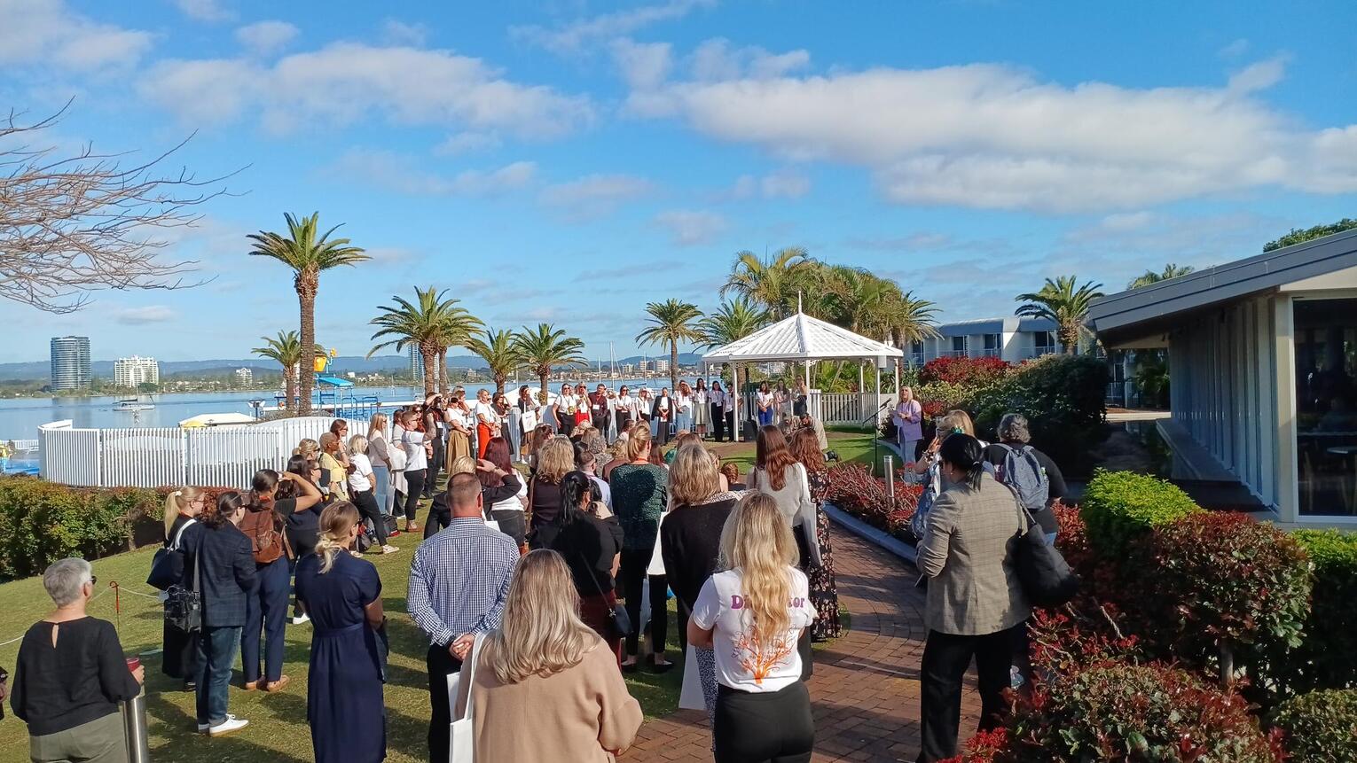 A large group of people gathers outdoors on a sunny day by the water, near palm trees and a white gazebo, with some people facing a speaker at the front. Modern buildings and blue sky are in the background.