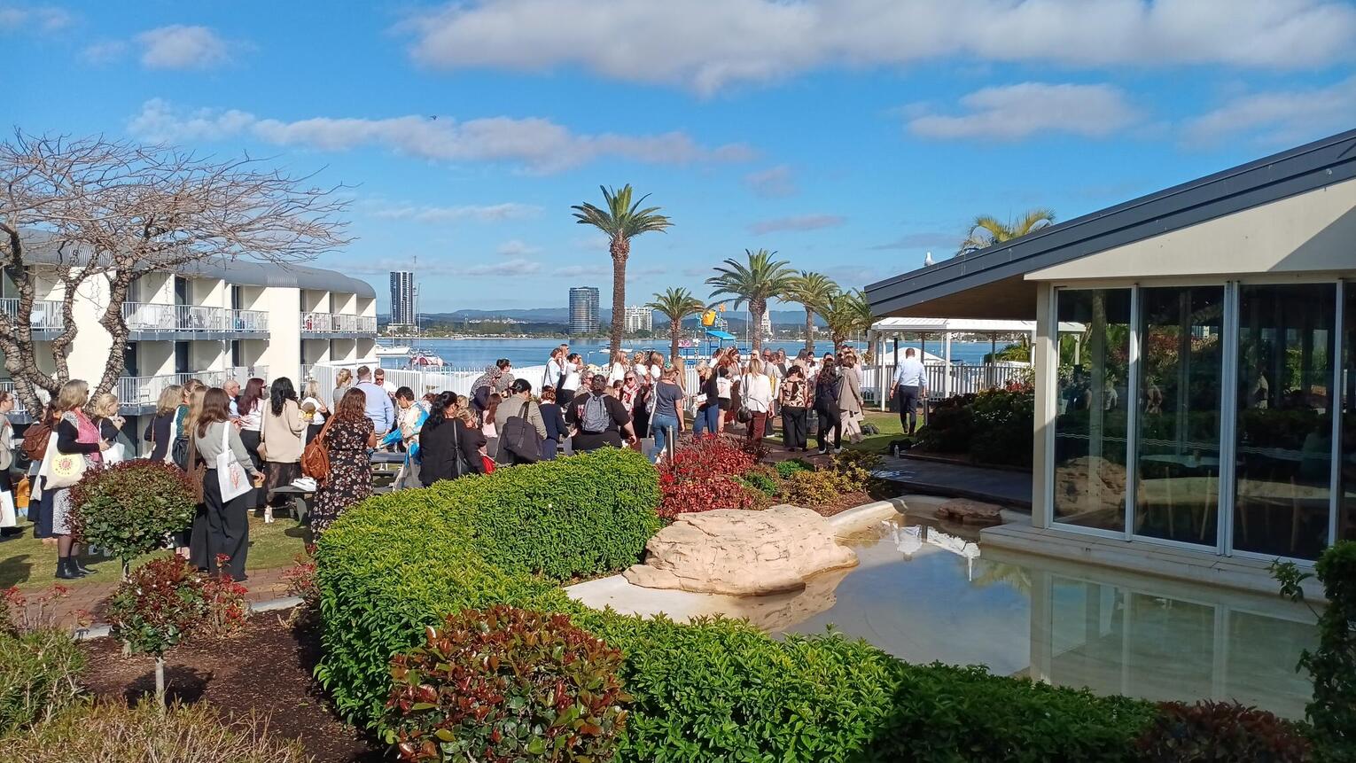 A large group of people gather outside near a hotel and a pond, surrounded by lush greenery, palm trees, and water views under a partly cloudy sky.