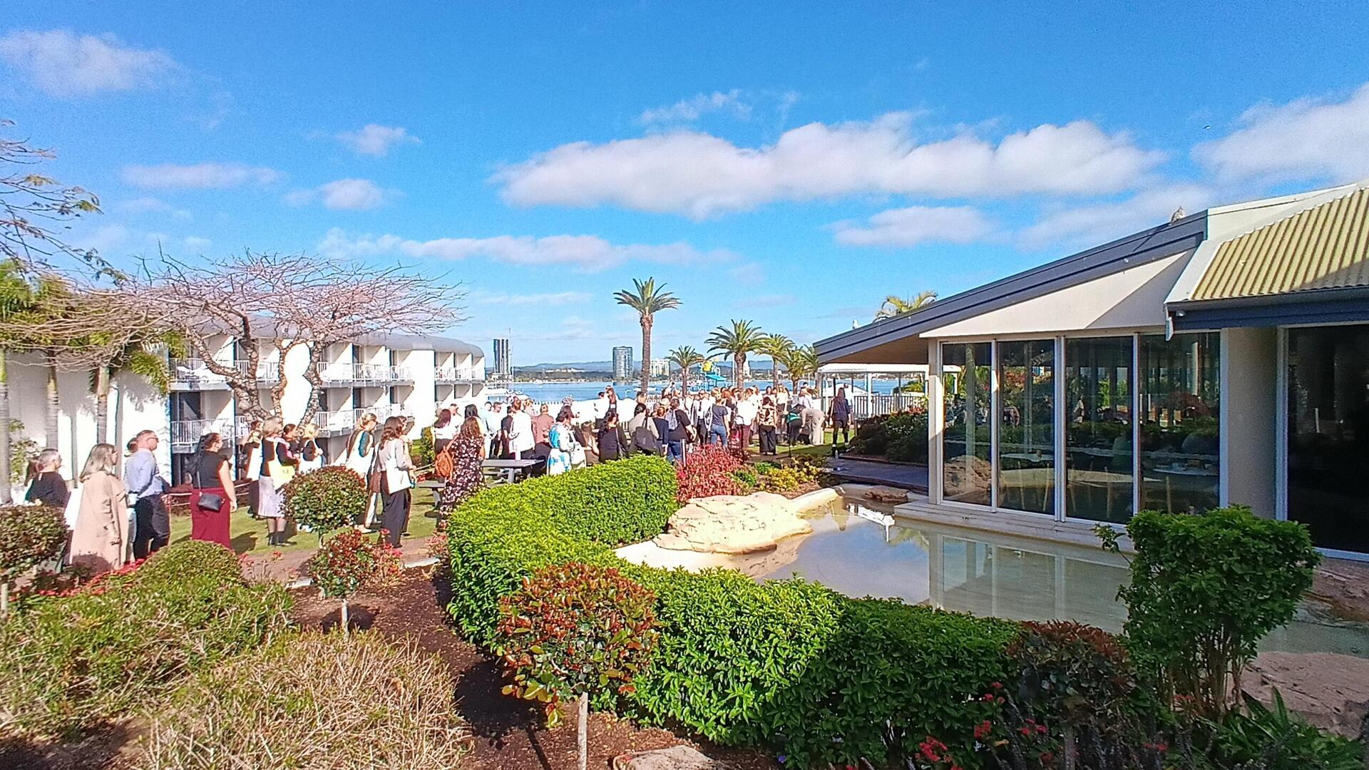 A large group of people gathers outdoors near a modern building with glass walls, landscaped gardens, and a small pond. Palm trees and blue sky with scattered clouds are in the background.