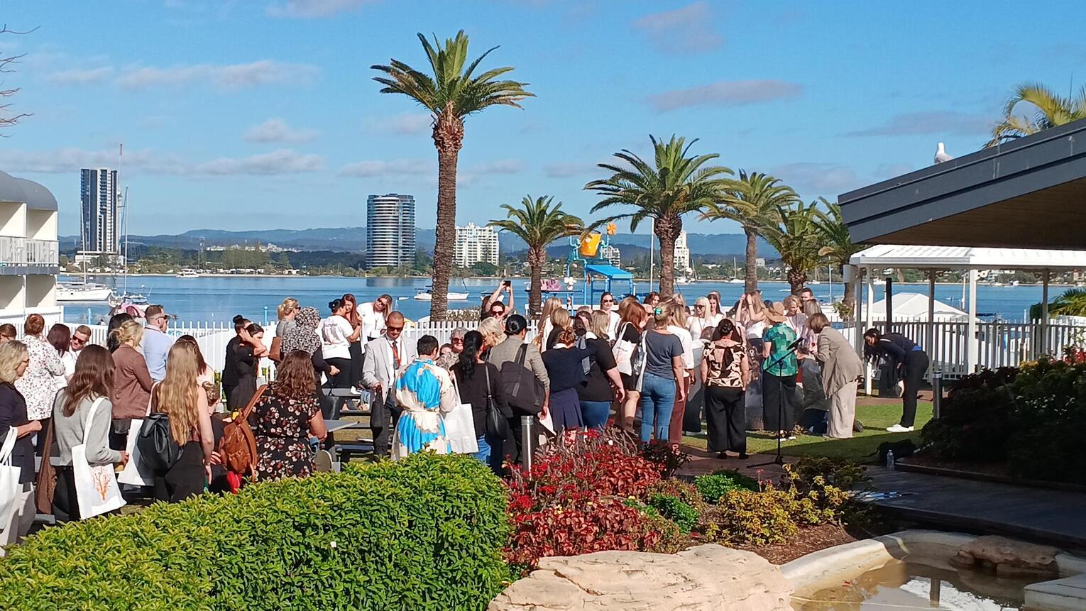 A large group of people gather outdoors near a waterfront, surrounded by palm trees and city buildings in the background on a sunny day. Some are talking while others face a speaker near a blue playground structure.