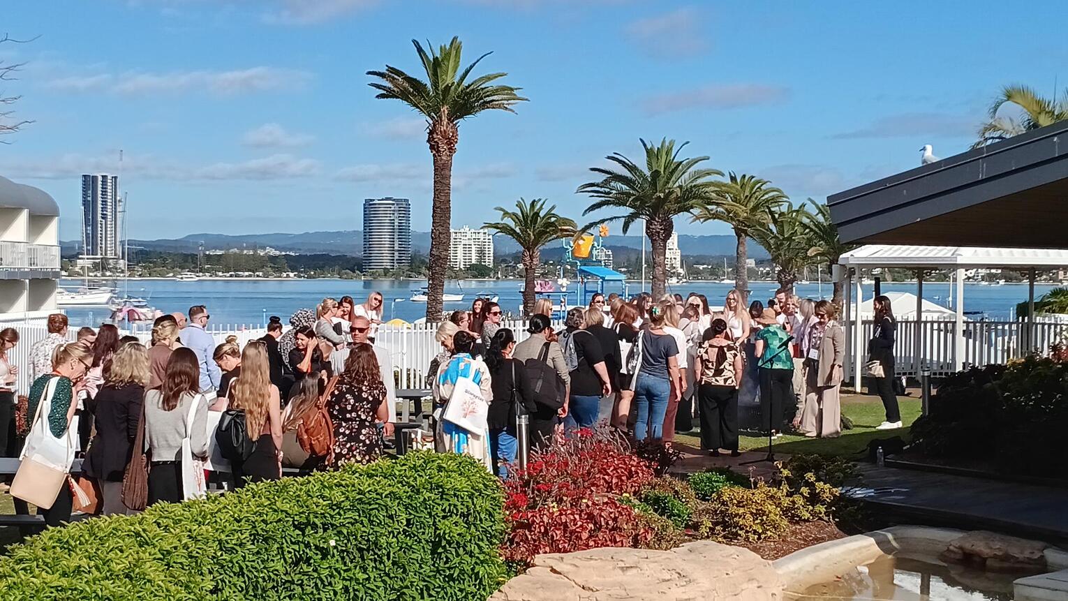 A large group of people gather outdoors near a waterfront, surrounded by palm trees and modern buildings under a blue sky, with some greenery and a white fence in the foreground.