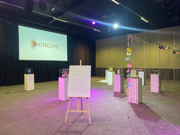 A gallery-style exhibition space with display pedestals, an easel with text, decorative hanging butterflies, and a large screen showing the Encore logo on the wall. The room is softly lit with purple accents.