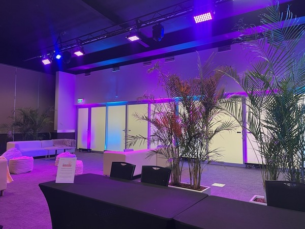 A modern lounge area with potted plants, sofas, and bean bags, illuminated by purple and blue lights. A black table with a sign stands in the foreground. The atmosphere is relaxed and inviting.