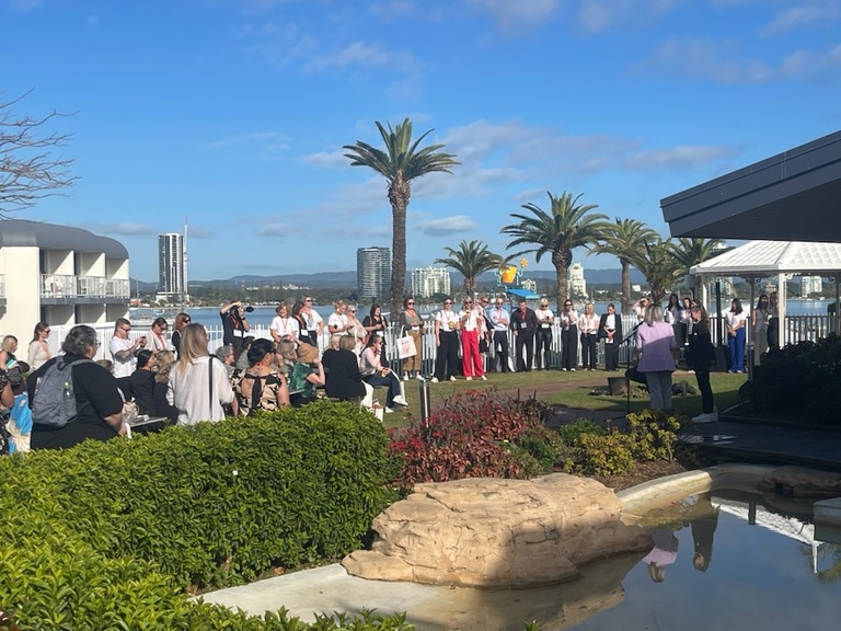 A large group of people gathers outdoors by a waterfront, near palm trees and a pond, listening to a speaker. Tall buildings and blue sky with clouds are visible in the background.