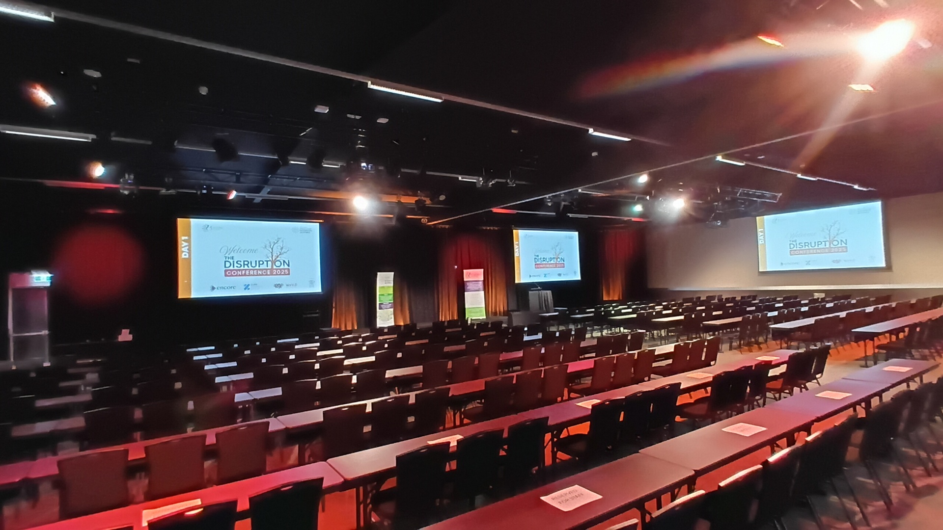 A large conference room with rows of empty tables and chairs, two projection screens displaying Disruption Conference 2023, and banners on stage under bright lights.