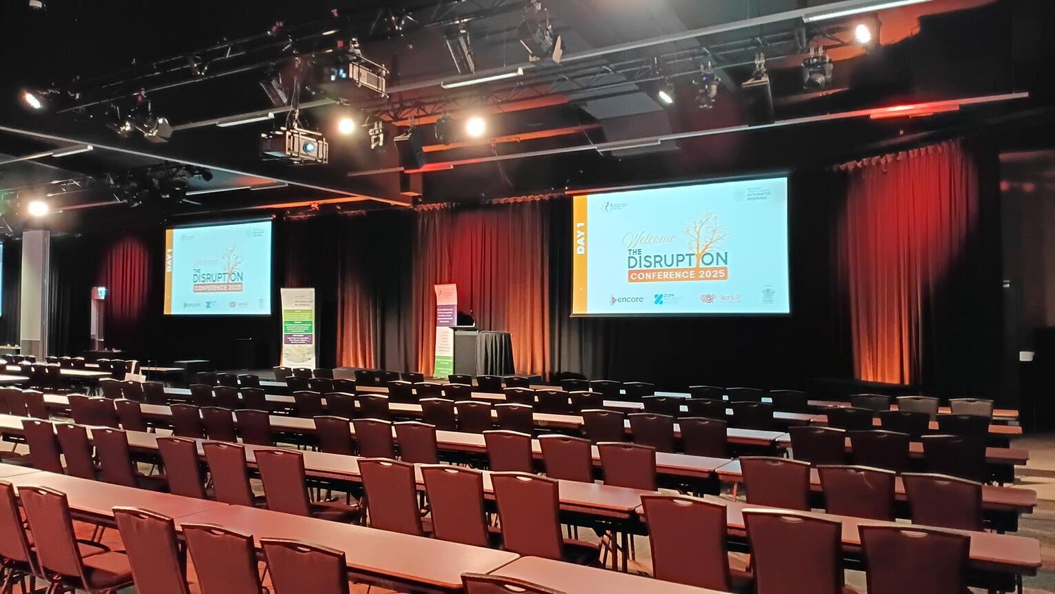 A large, empty conference room with rows of chairs and tables facing a stage, two projection screens displaying DISRUPTION CONFERENCE 2023, and reddish-orange curtain backdrops.