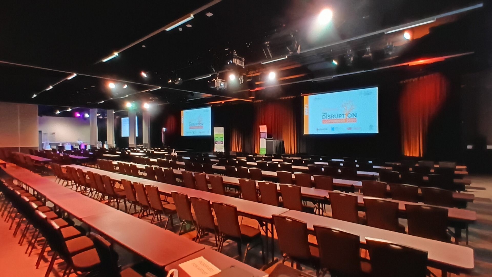 A large, empty conference room with rows of chairs and tables facing two projection screens displaying a Disruption event banner. The room is dimly lit with colored lighting.