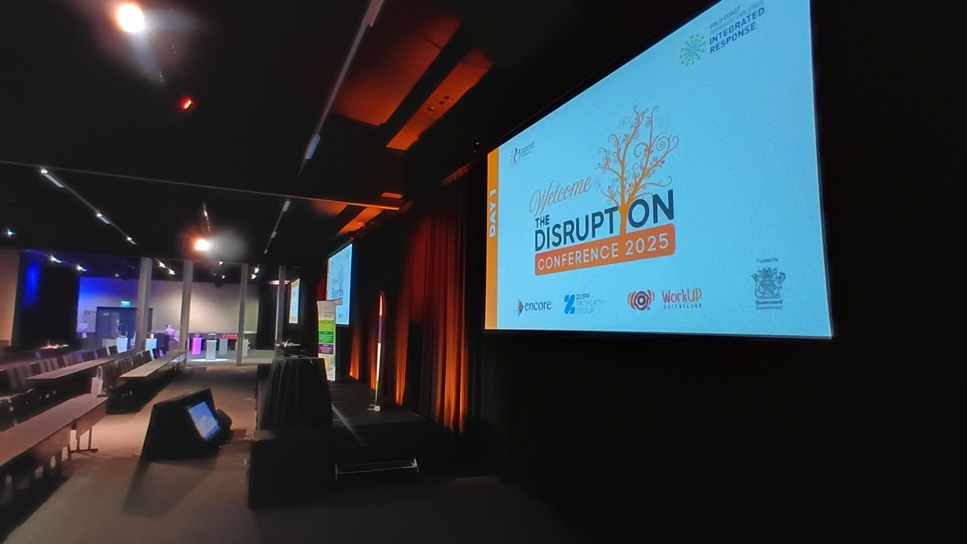 A large conference room with rows of empty chairs faces a big screen displaying Welcome to the Disruption Conference 2025 and sponsor logos, with colorful lighting along the walls.