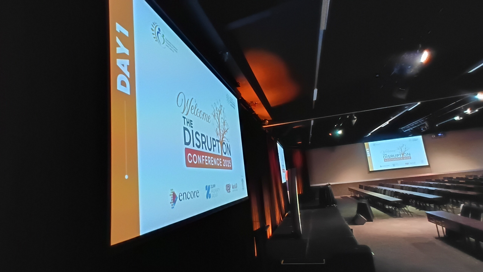 A large screen in a dimly lit conference room displays “Welcome to the Disruption Conference 2023, Day 1.” Rows of empty chairs and another screen are visible in the background.