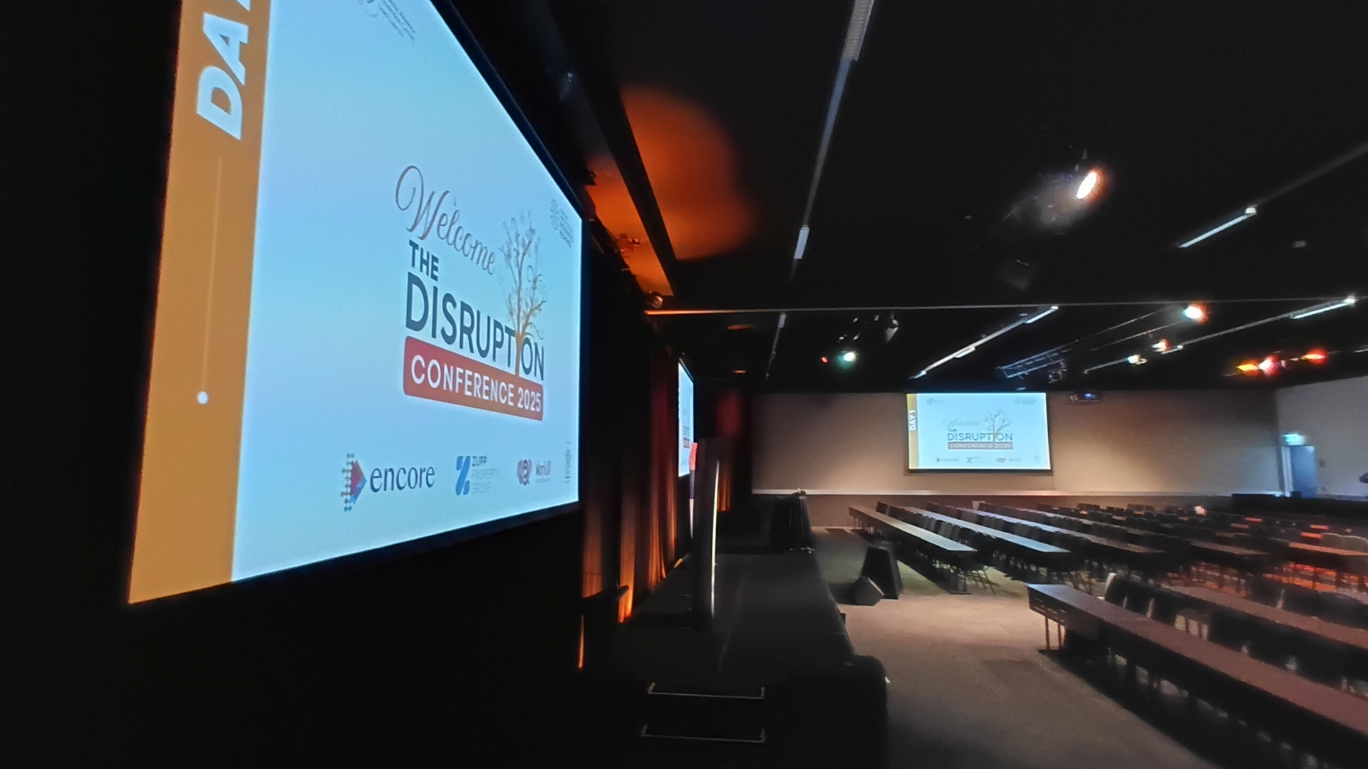 A conference room with long tables and chairs faces a large screen displaying “Welcome to the Disruption Conference 2023” with sponsor logos. The room is dimly lit, and a similar screen appears at the far end. Encore Case Study: Domestic Violence Prevention Centre . Sea World Gold Coast