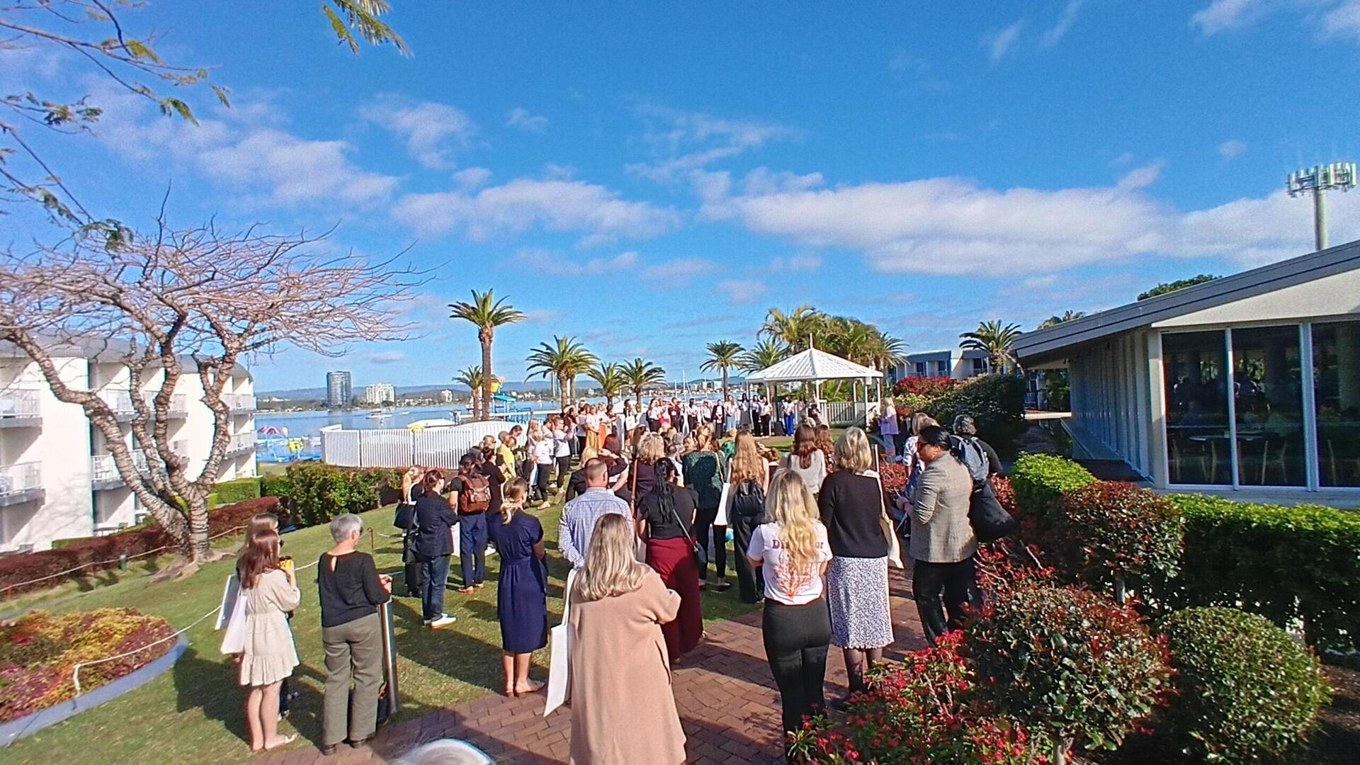 A large group of people dressed formally gather outdoors for an event near water, with palm trees, gardens, and buildings under a bright blue sky with scattered clouds.