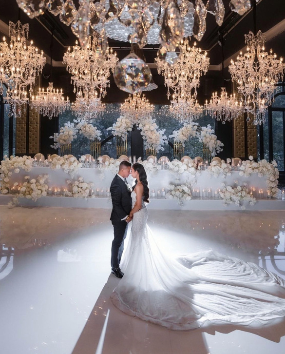 A bride and groom share a romantic moment on a glossy white floor beneath sparkling chandeliers, surrounded by elegant white floral arrangements and soft candlelight in a lavish weddings setting.