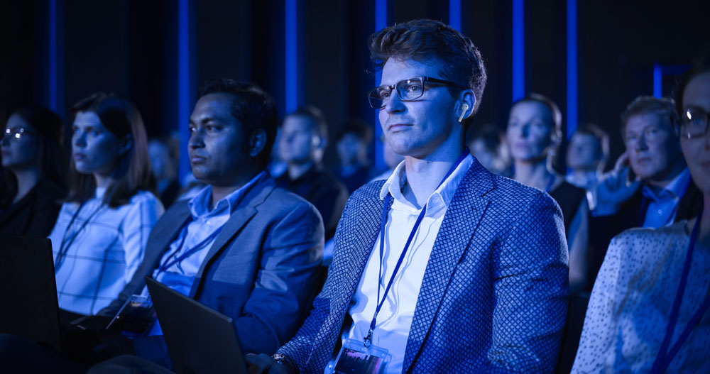 A diverse group of people sits attentively in an audience at a professional conference or seminar. Most wear business attire and name badges; some have laptops. The room is dimly lit with blue lighting.
