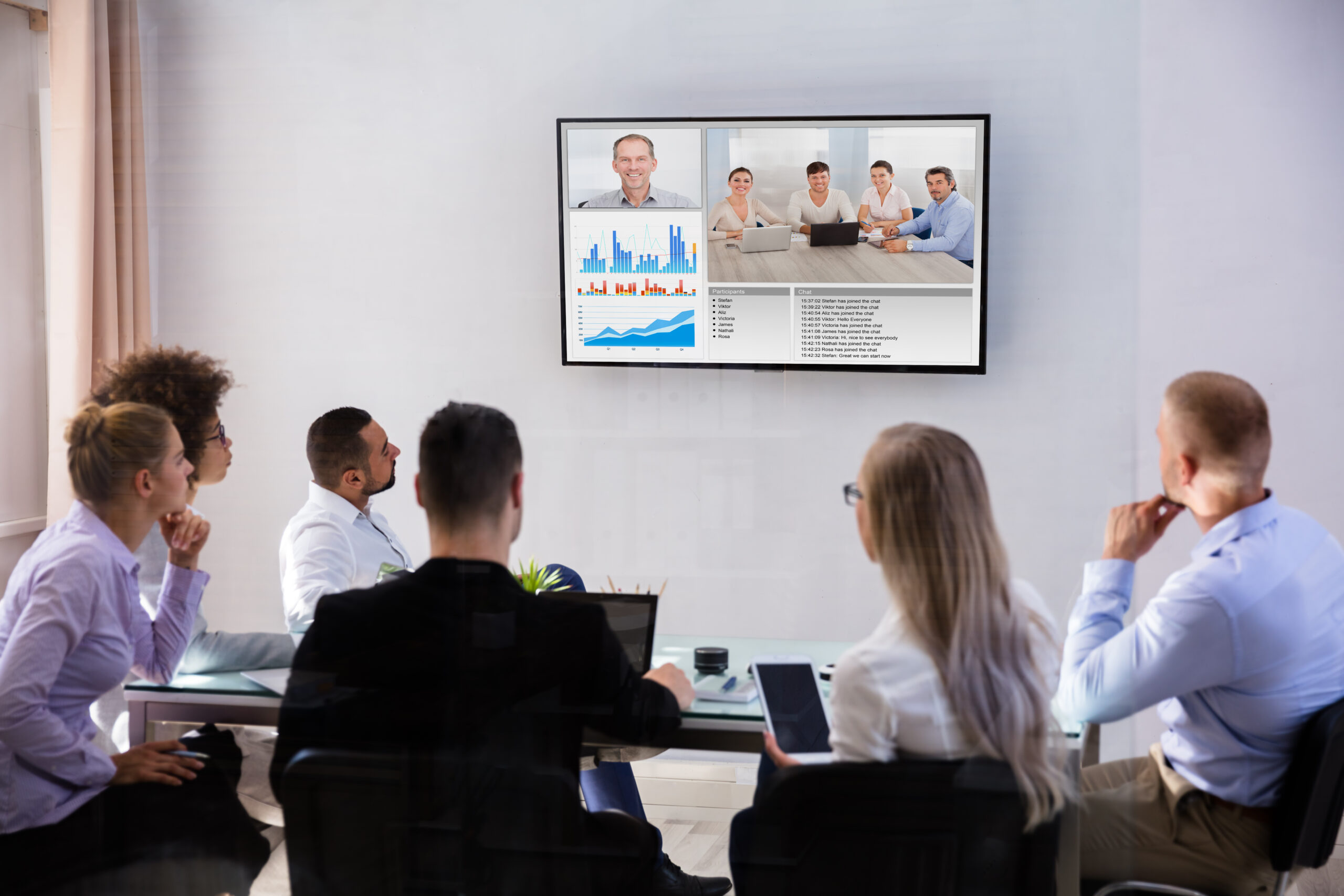 Six people sit around a glass table in an office, watching a video conference displayed on a wall-mounted screen showing charts and participants in a remote meeting.