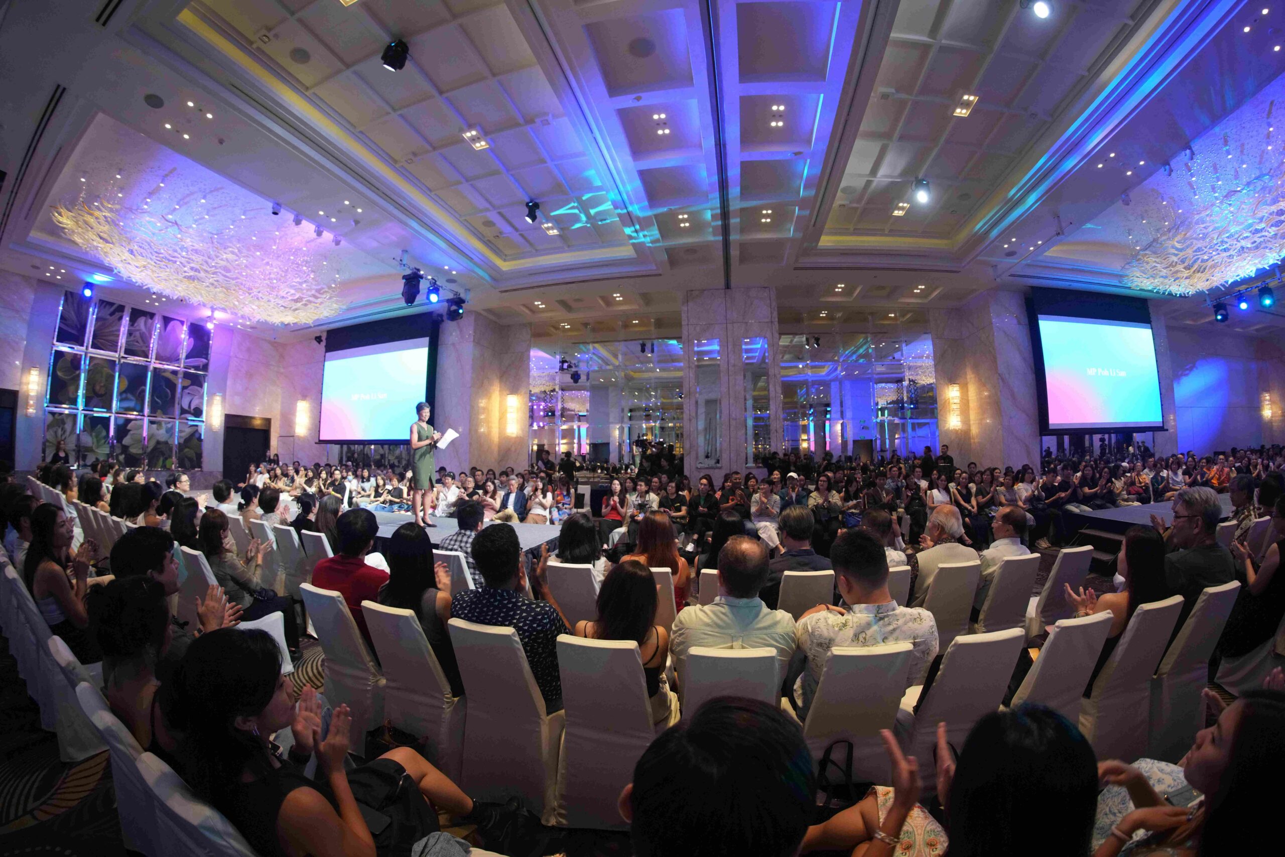 A large audience sits in a grand, brightly lit ballroom with chandeliers, watching a Fashion for Cancer Singapore speaker on stage. Two large screens display text on either side. The atmosphere is lively and formal.