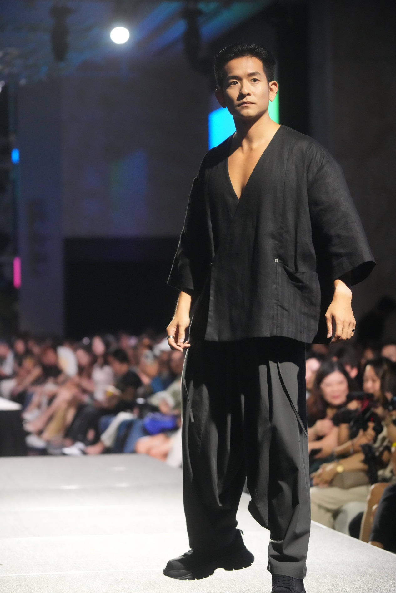 A male model walks down a runway at Fashion for Cancer Singapore, wearing a loose-fitting, black V-neck top and wide black pants, while an audience watches in the background.