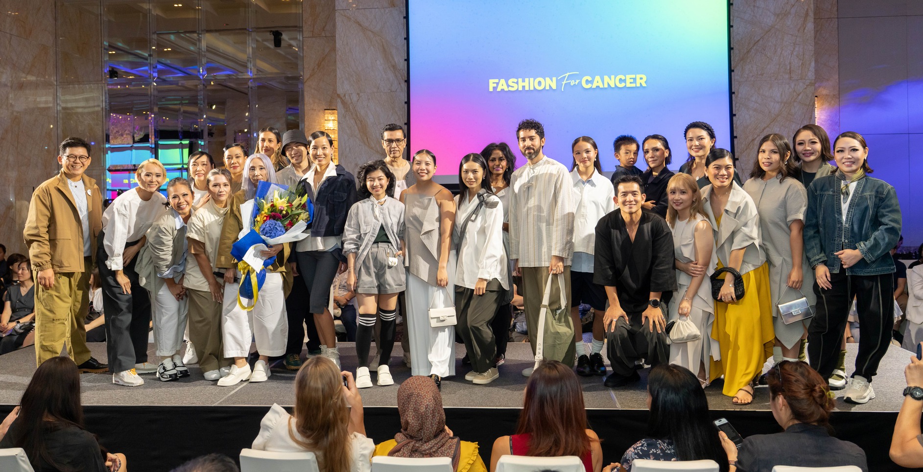 A large group of people, many dressed in stylish outfits, stand together on a stage in front of a screen that reads Fashion for Cancer Singapore, while an audience watches and some people hold flowers.