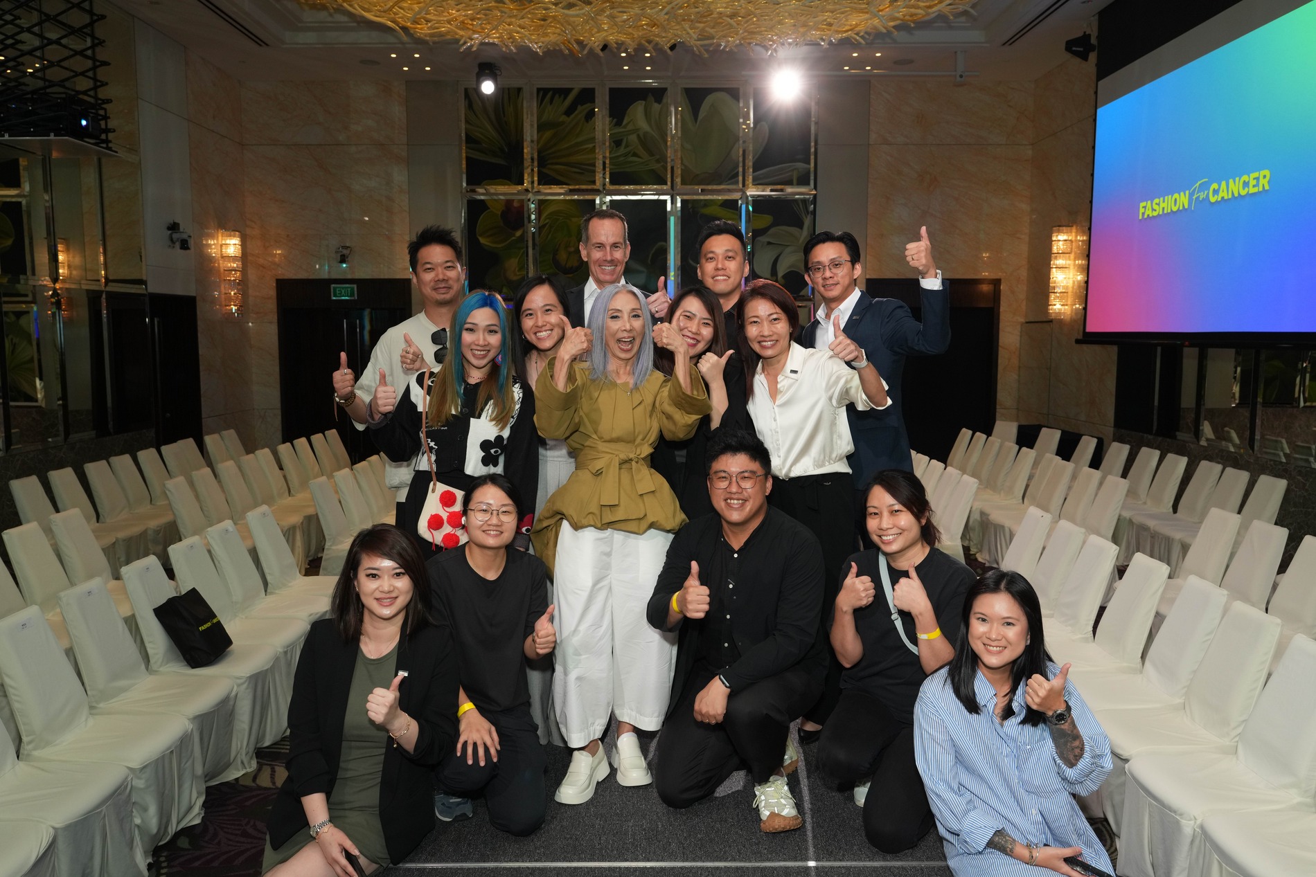 A group of people stand and kneel in two rows, smiling and giving thumbs-up gestures in a well-lit event space for Fashion for Cancer Singapore, with empty rows of white chairs and a screen displaying “FASHION x CANCER.”.