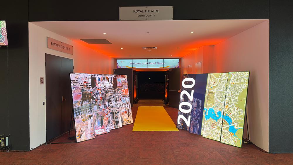 Large digital screens showing aerial maps and 2020 frame a yellow carpet entrance to the Royal Theatre, with a welcoming sign in the background and warm orange lighting.