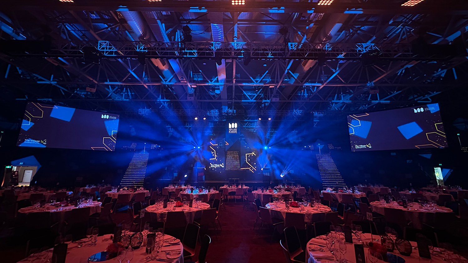A large event hall set up for a formal dinner, with round tables and chairs arranged for guests. Blue stage lights illuminate the stage and ceiling, and two large screens display graphics on either side of the stage.