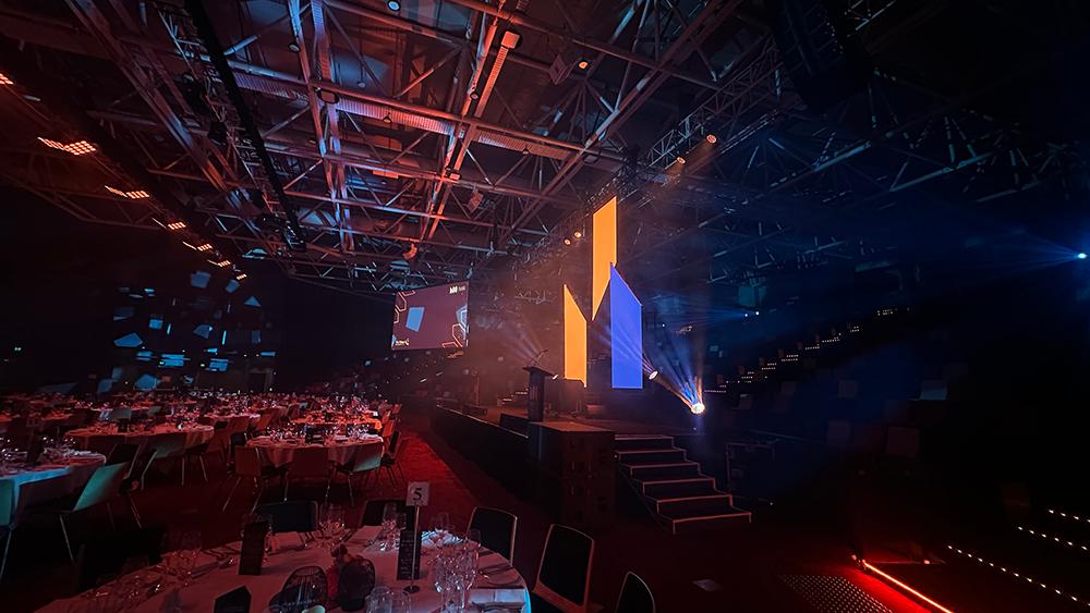 A dimly lit event hall with round tables set for a formal gathering, featuring a large stage with colorful vertical LED screens and dramatic blue and orange lighting.