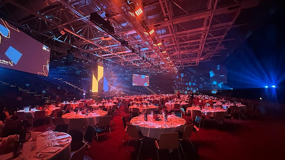 A large banquet hall with round tables set for dinner, dramatic stage lighting in blue and orange, and screens displaying abstract visuals on the walls, creating a vibrant, event-like atmosphere.