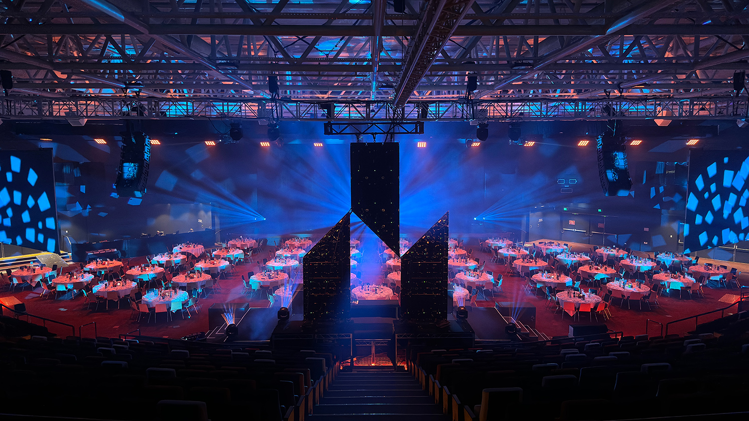 Large banquet hall set up for an event, with round tables covered in white cloths, dramatic blue and pink stage lighting, and abstract stage decorations in the center. Rows of empty seats face the tables and stage.