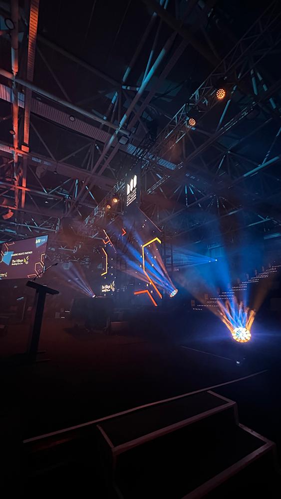 A dark indoor stage with colorful blue and orange spotlights, geometric LED lights, and a large truss structure. A podium is on the left and illuminated stairs lead up to the stage. The ceiling has exposed beams.