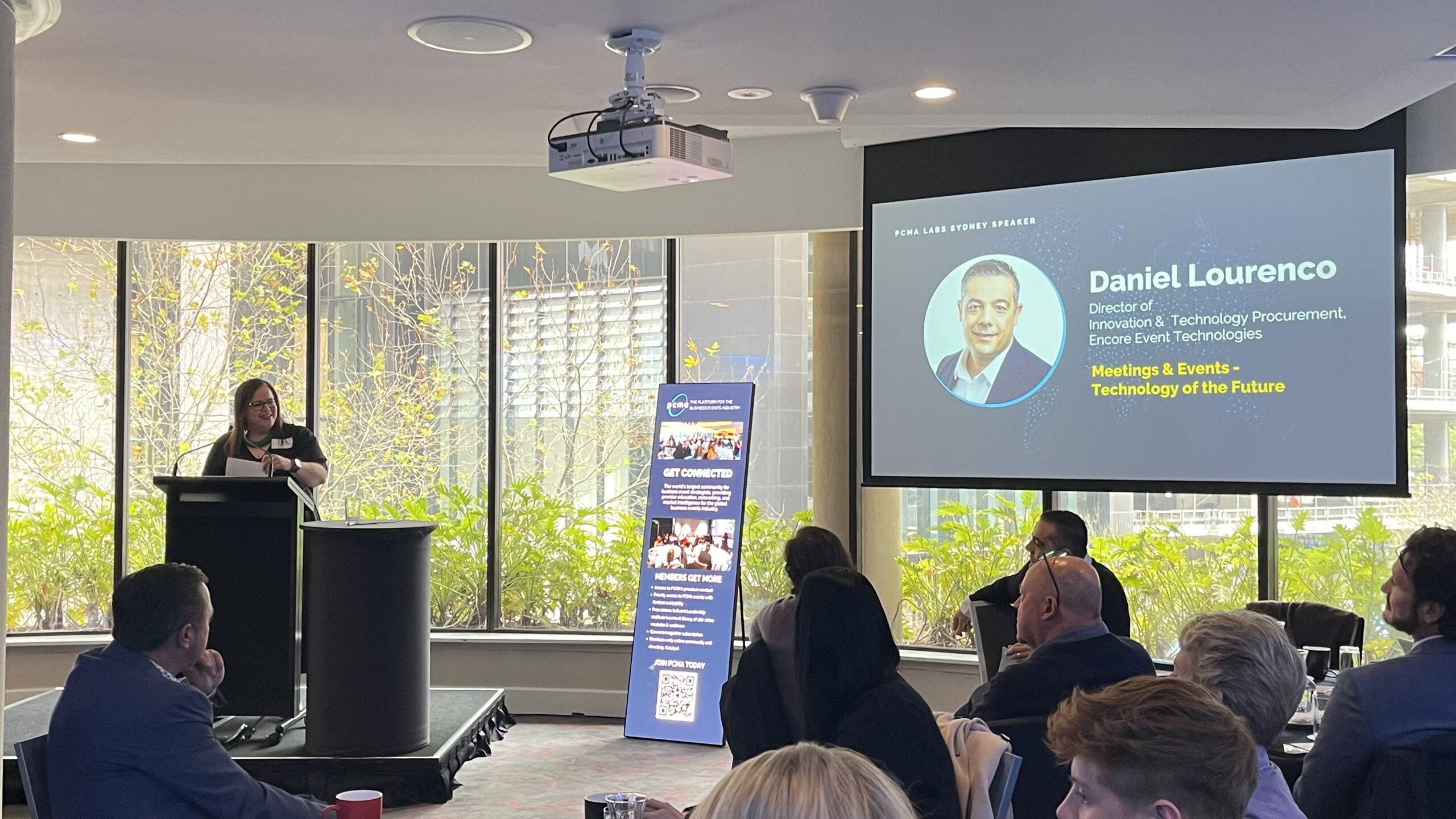 A woman speaks at a podium in a conference room as attendees listen. A screen displays information about a guest speaker, Daniel Lourenco, and his talk on Technology of the Future. Large windows provide natural light.