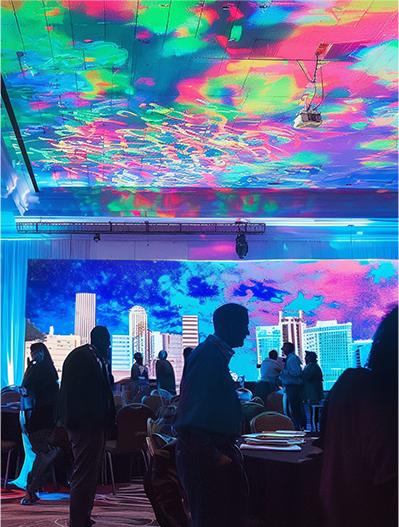 People stand and mingle in a large room with colorful, abstract lights projected on the ceiling and a city skyline image displayed on a screen—showcasing optional event enhancements that elevate the atmosphere.