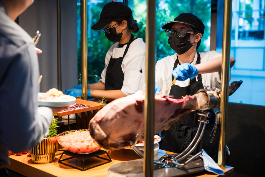 Two people in black caps and masks carve a large leg of cured meat at a buffet station, preparing a plate of food for a customer. The scene is indoors with greenery visible outside the window.