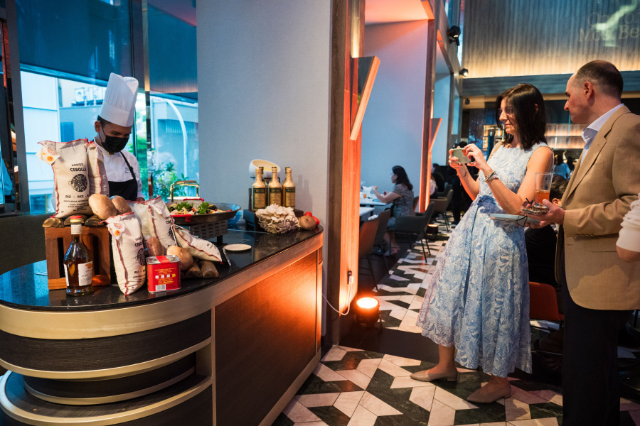A chef in uniform prepares food at a counter with ingredients, while a woman in a blue dress takes a photo and a man beside her holds a plate and wine glass in a stylish restaurant.