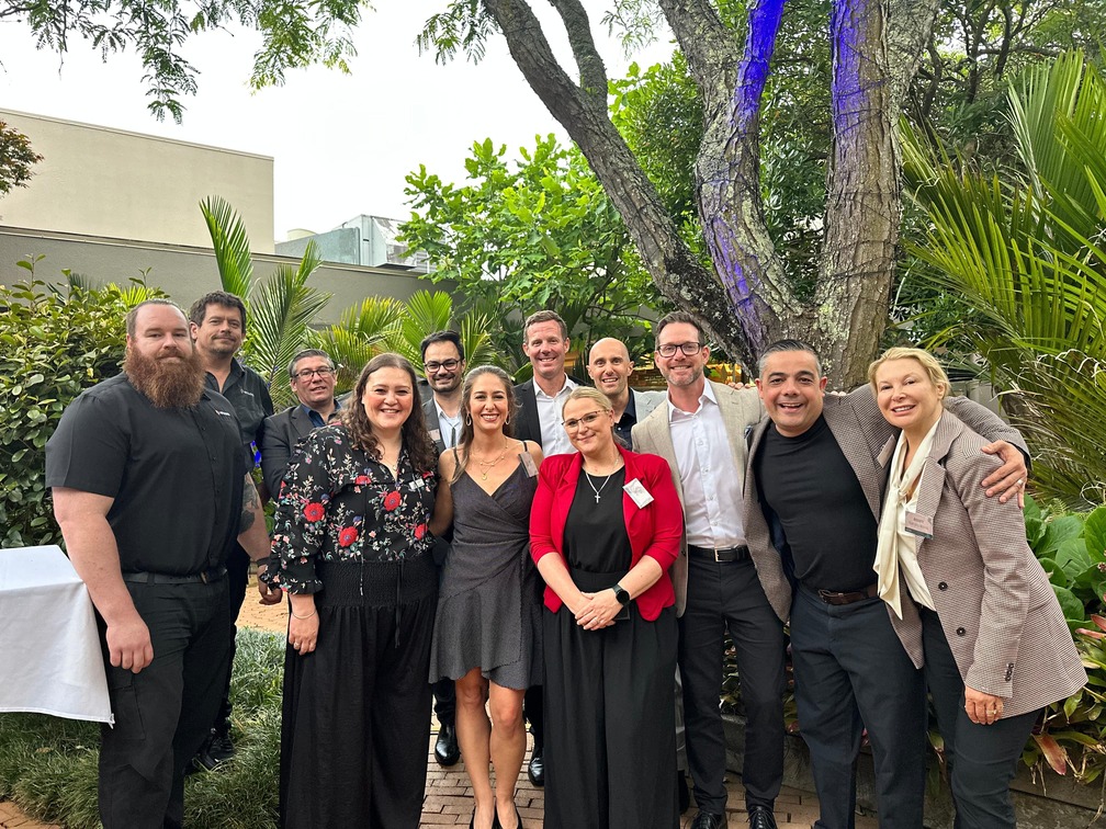 A group of twelve adults, dressed in semi-formal and business attire, smile for a group photo outdoors in a lush garden with green plants and trees in the background |Waipuna Hotel and Conference Centre - Strategic Partnership with Encore - Encore partners with Waipuna Hotel and Conference Centre