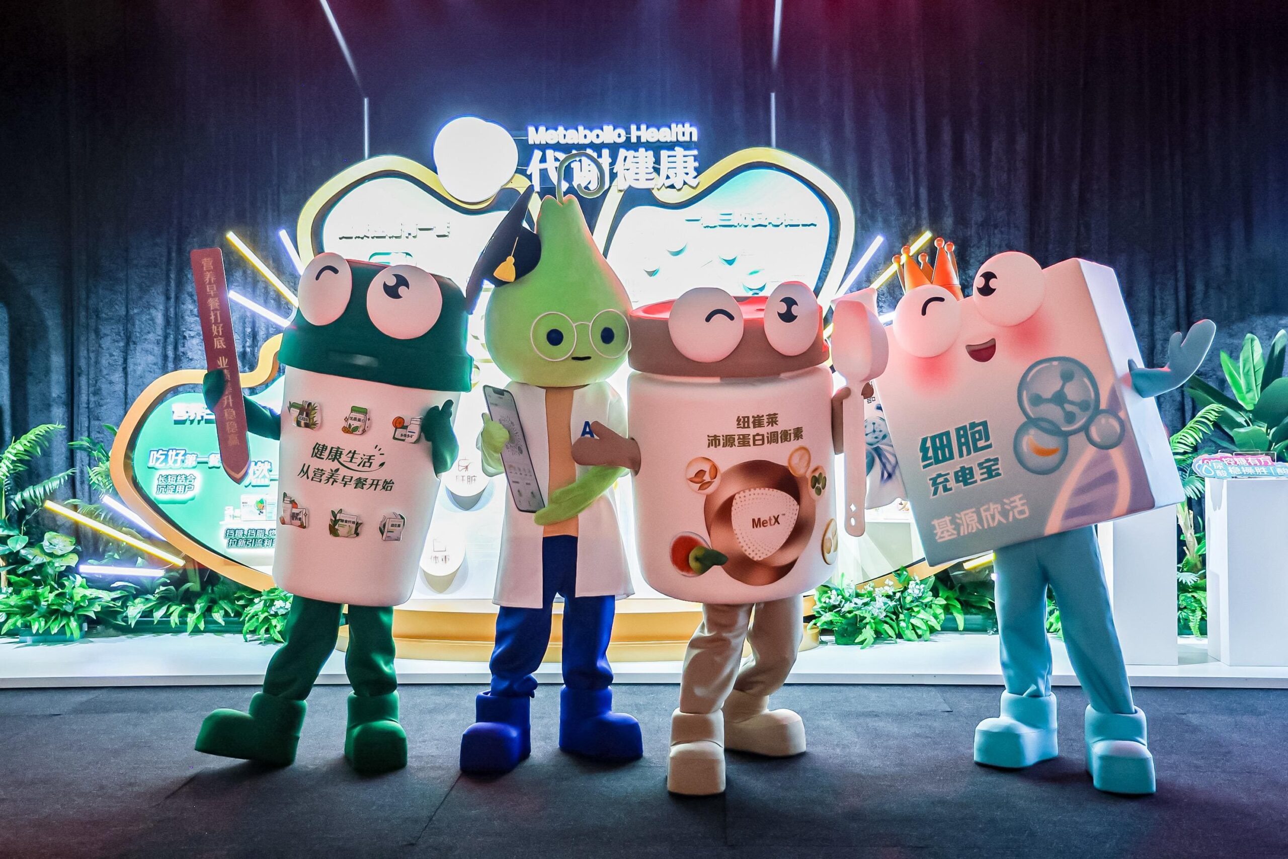 Four people in colorful, cartoon-style costumes pose together at a brightly-lit health event, possibly celebrating product launches. Chinese text and plants fill the background, while the playful costumes feature large props related to health or wellness. Encore creates brand activations and product launches excite and engage.