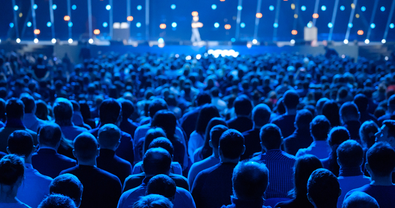 A large audience sits in a dimly lit auditorium facing a brightly lit stage, where a single person stands speaking. Blue lighting creates a dramatic atmosphere, and strings of lights hang behind the stage. Will AI replace event planners in future?
