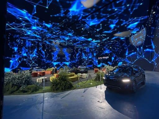 A futuristic indoor space with a ceiling covered in glowing blue digital network patterns hosts product launches, featuring various seating areas, greenery, and a black car on display. People are gathered near the seating, and the name Amy Shah is visible. Encore creates brand activations and product launches excite and engage.