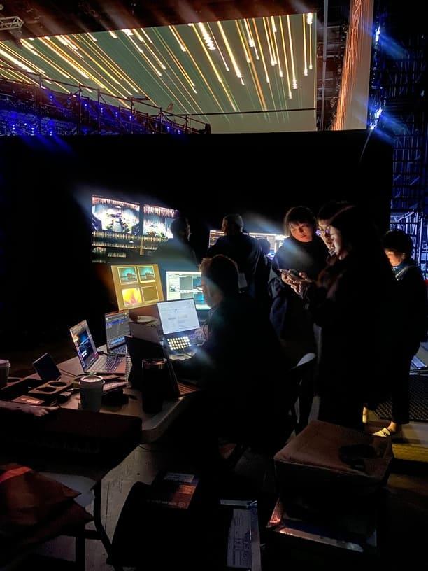 Several people work at a table with laptops and monitors in a dark backstage area, illuminated by screens. A large display with streaks of light hints at a live event or product launches happening behind the scenes. Encore creates brand activations and product launches excite and engage.