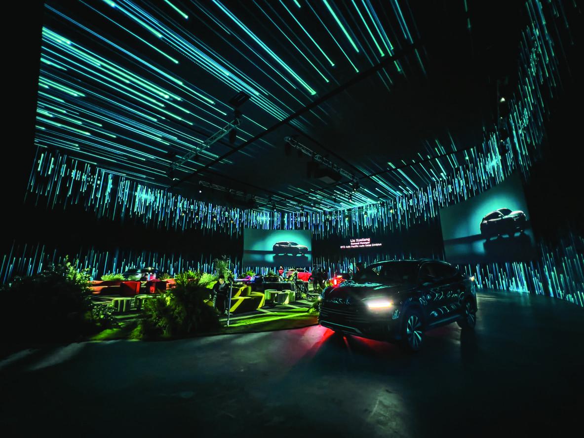 A dark, futuristic room with glowing blue lines on the walls and ceiling showcases a black car in the foreground—illuminated seating areas enhance the high-tech, immersive atmosphere, perfect for cutting-edge product launches. Encore creates brand activations and product launches excite and engage.