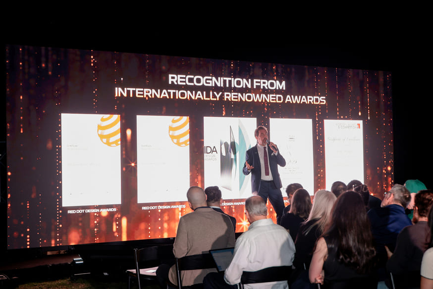 A speaker addresses an audience in front of a large screen displaying Recognition from Internationally Renowned Awards and several award images and text during product launches at an indoor event. Encore creates brand activations and product launches excite and engage.