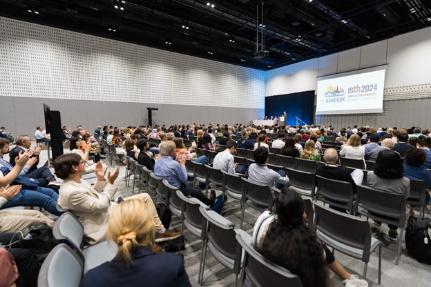 A large audience sits in a modern conference hall, facing a stage with a presentation screen displaying ISTH 2024 Bangkok. Some people are clapping while speakers, including special guests from UC Berkley - Light the Way, stand at the front.