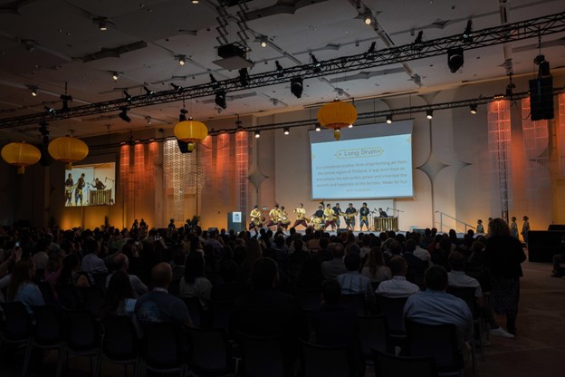 A large audience watches a group of performers on stage in a spacious, modern auditorium. Warm lighting and hanging lanterns decorate the ceiling as the UC Berkeley - Light the Way presentation text shines on the screen behind them.