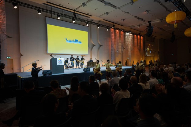 A group of performers in traditional attire plays instruments on stage at a large indoor UC Berkeley – Light the Way event, with an audience watching. The stage features a yellow screen and decorative lighting panels.