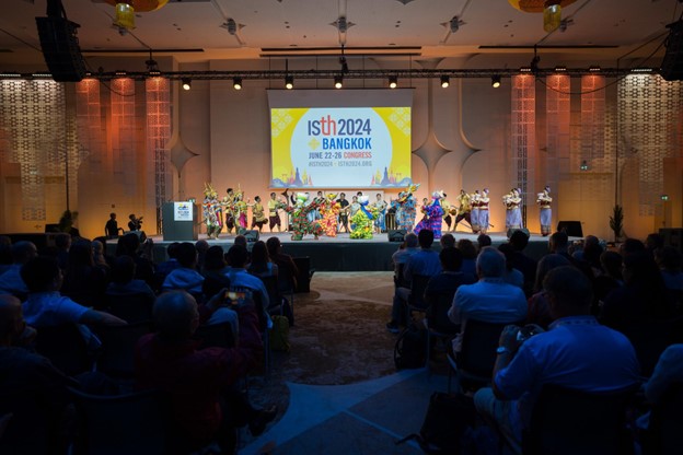 A large audience watches a colorful traditional dance performance on stage at the ISTH 2024 Bangkok Congress, with a bright event banner and a nod to UC Berkley - Light the Way displayed in the background.