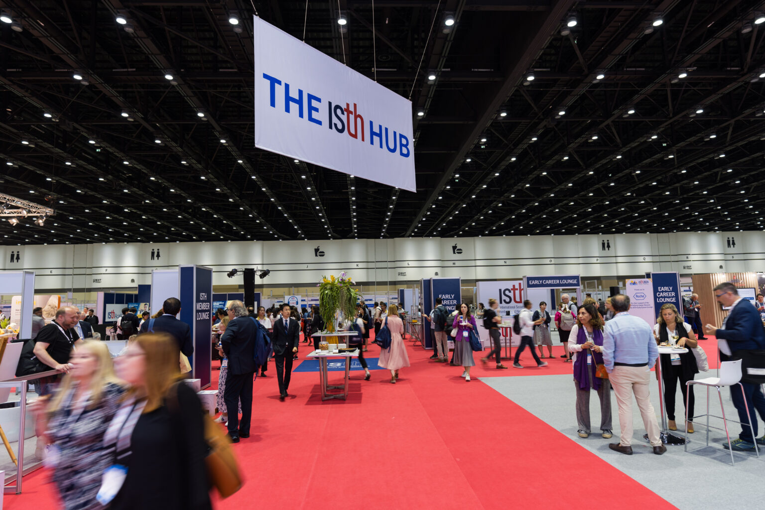 A busy convention hall with people walking and talking on a red carpet. A large sign overhead reads “THE ISth HUB,” while UC Berkley - Light the Way booths and displays are visible in the background.