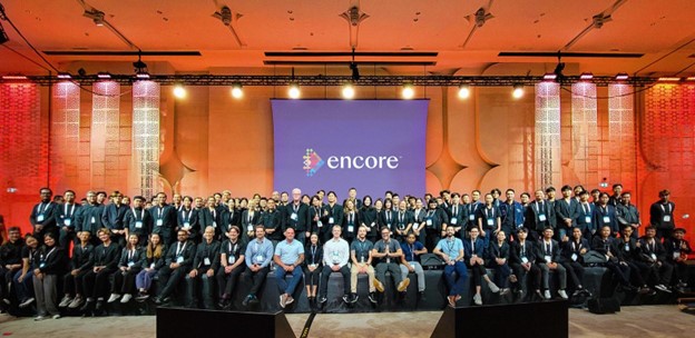 A large group of people in business attire pose together on stage in front of a screen displaying the Encore logo at a corporate event inspired by UC Berkeley – Light the Way, held in a spacious, well-lit hall.